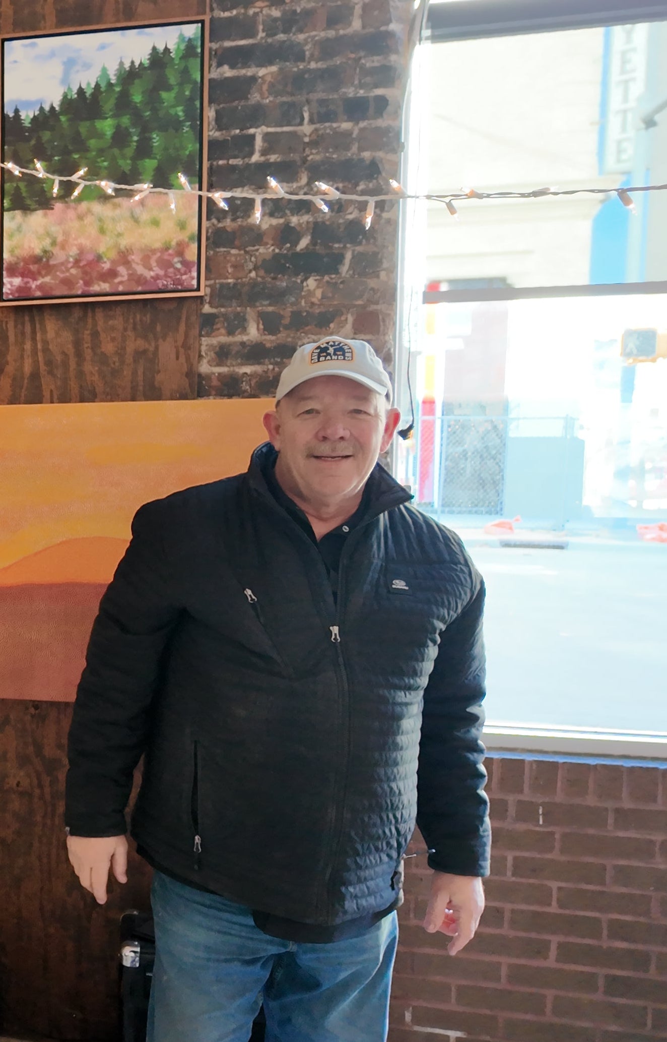 A man wearing a black quilted jacket, blue jeans, and a white baseball cap stands in front of a brick wall and window in an indoor setting.